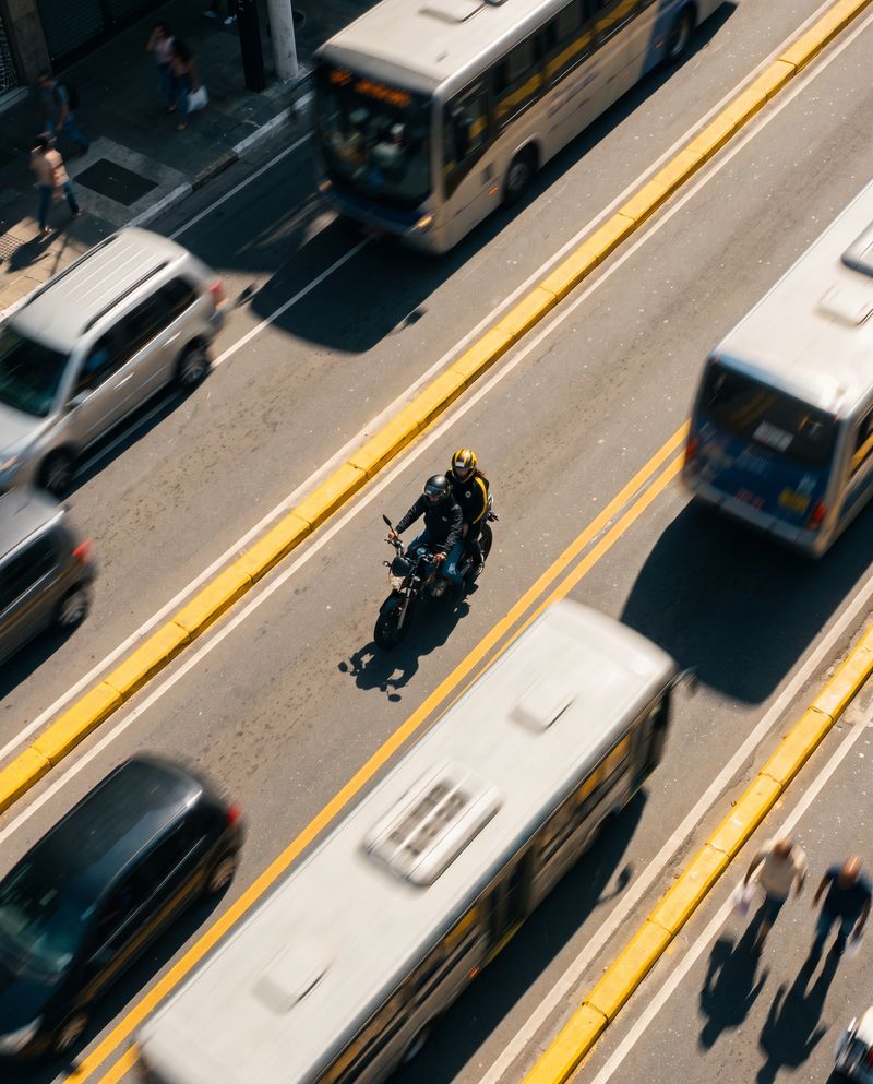 Vista do alto de uma moto passando entre ônibus e carros em uma avenida