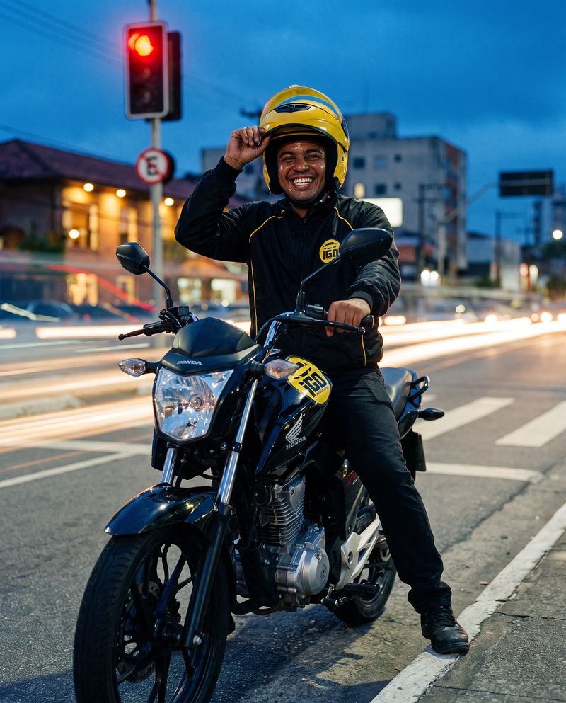 Piloto da iGO Motos sorrindo e cumprimentando com a mão no capacete, parado na moto ao anoitecer